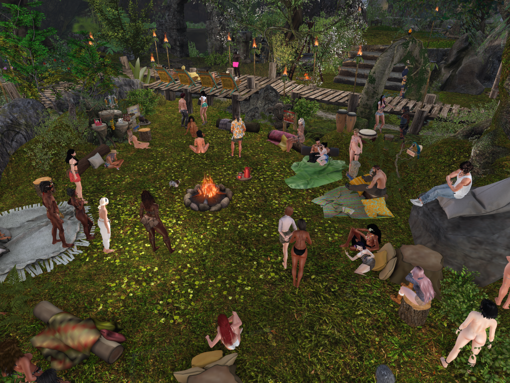 People in varied attire gather around a fire in the jungle camp, sitting and standing, surrounded by nature